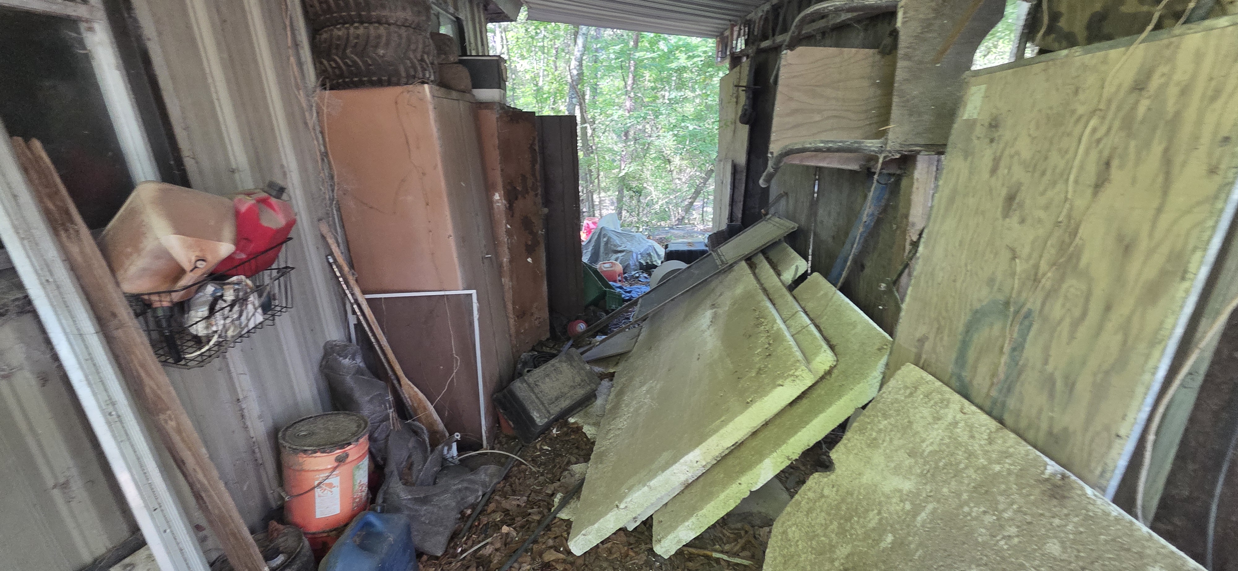 Cluttered shed and covered structure before cleanout — plywood, cans, and mixed junk — Go Forward Haulers