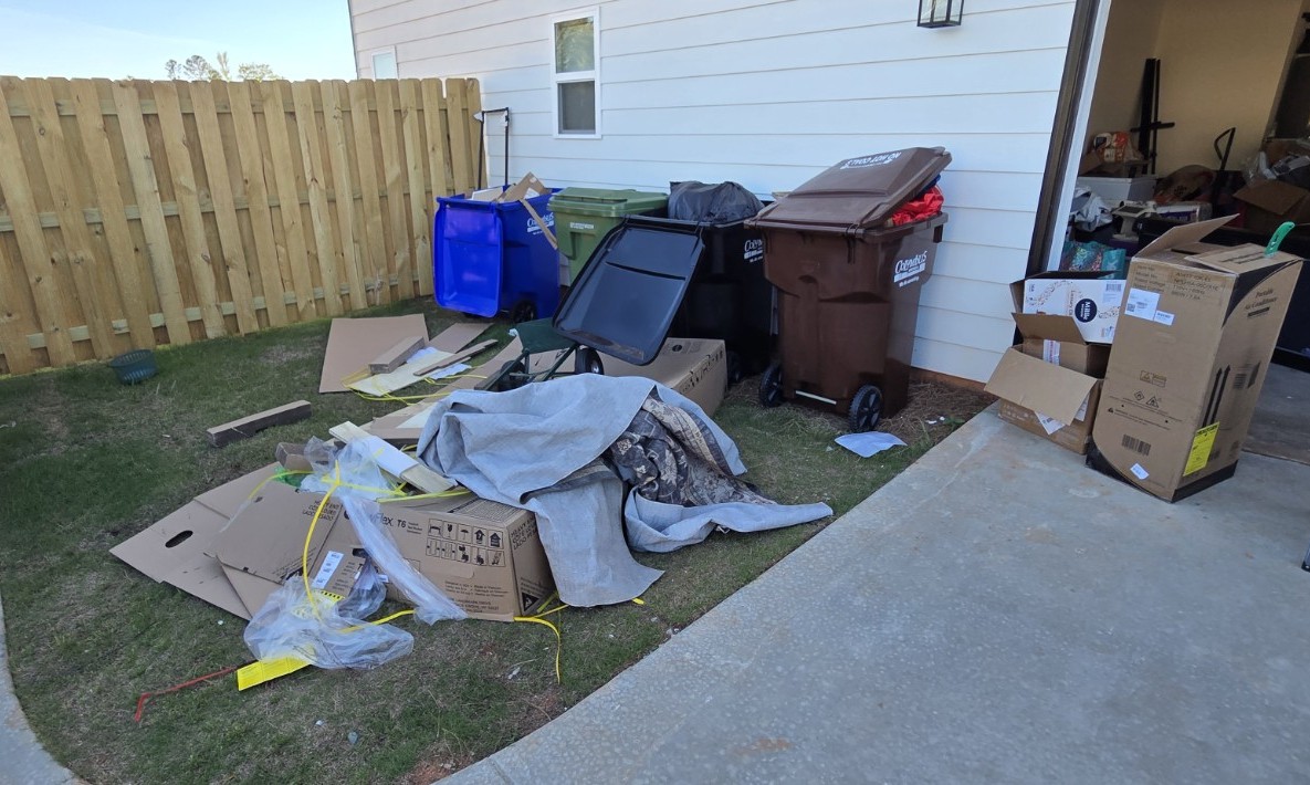 Yard junk and debris pile before removal in Midland GA — cardboard, tarp, and bulk items at fence
